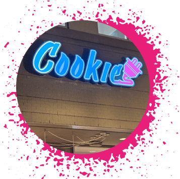 Blue, pink, and purple gradient sign says 'Cookie Plug' over a tan storefront with a pink paint splatter circular border.
