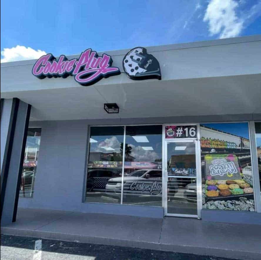 Pink, white, and black sign over a storefront that says 'Cookie Plug' with a black, white, and gray logo with a cookie and a plug attached to it. The windows have large pictures of cookies in the window in a box that says 'it was all a dream' in graffiti letters.