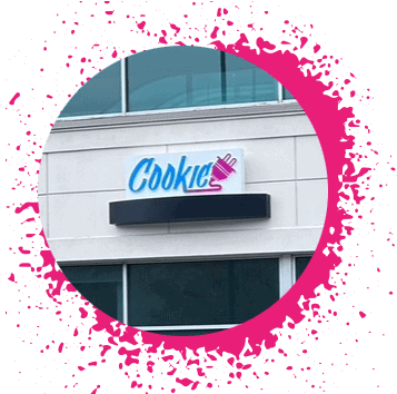 Blue, pink, and purple gradient sign says 'Cookie Plug' over a white brick storefront with large windows with a hot pink spray paint border