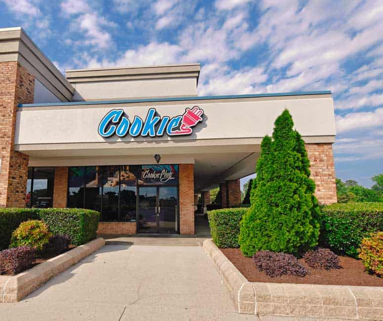 Tan and brick storefront that has a large sign that says 'Cookie' with an electrical plug attached to the end in a blue, purple, and pink gradient with pictures of large cookies in the window.