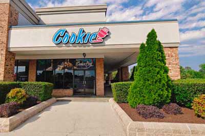 Tan and brick storefront that has a large sign that says 'Cookie' with an electrical plug attached to the end in a blue, purple, and pink gradient with pictures of large cookies in the window.