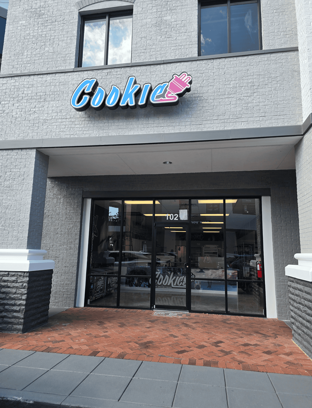 Gray brick storefront that has a blue and pink sign that says 'Cookie' with an electrical plug attached to the end of it.