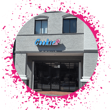 Gray brick storefront that has a blue and pink sign that says 'Cookie' with an electrical plug attached to the end of it.