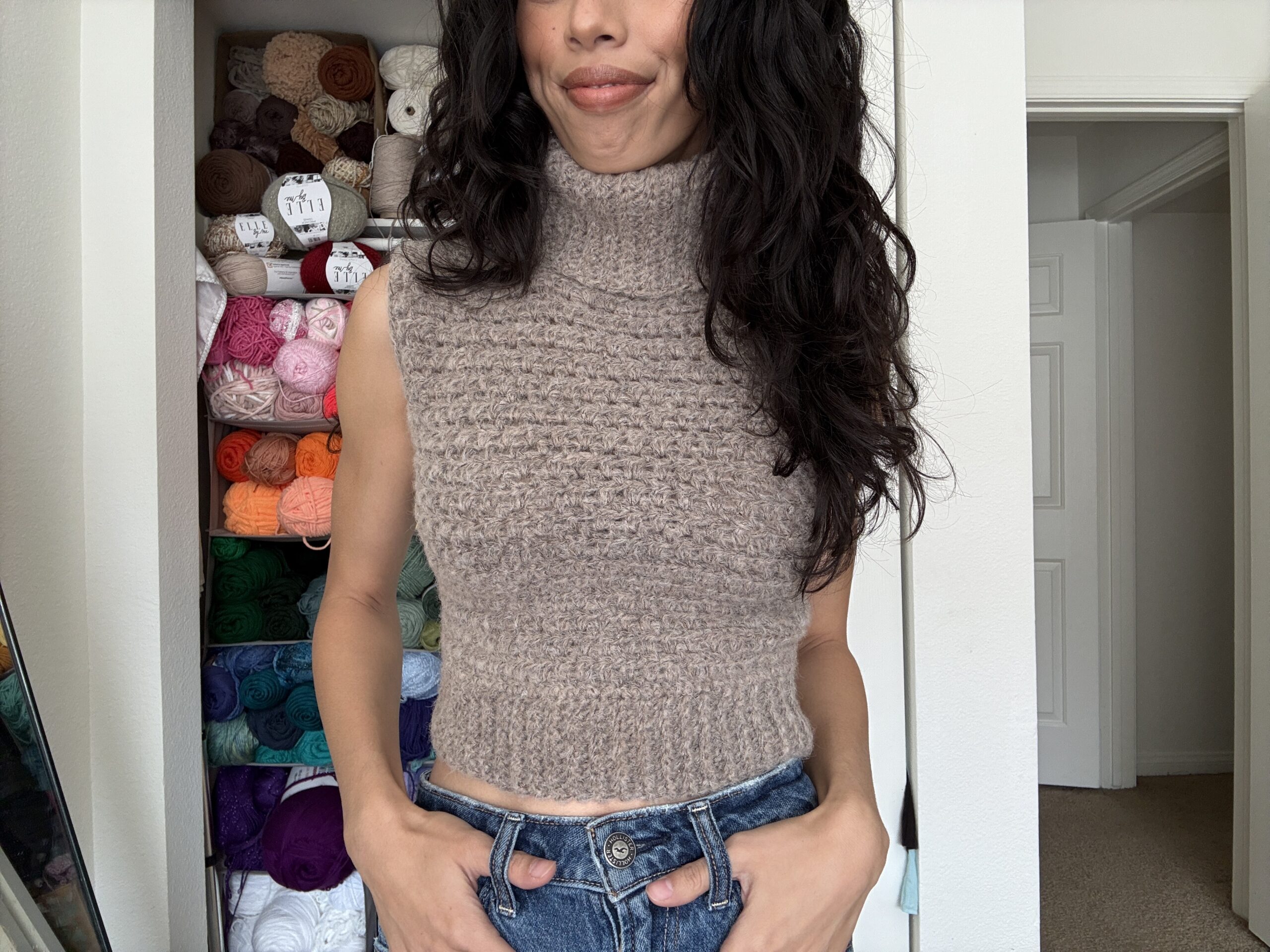Handmade sleeveless crochet turtleneck sweater in taupe yarn, styled with high-waisted jeans, in front of a colorful yarn shelf