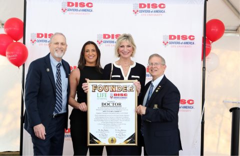 Four people stand in front of a DISC Centers of America banner, with two holding a framed Founder Life University certificate. Red balloons are visible in the background.
