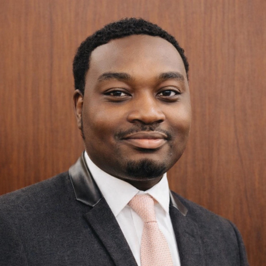 Man in a suit and light pink tie stands in front of a wood-paneled background, smiling slightly at the camera.