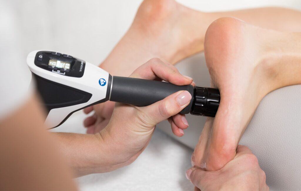 A person receives a medical treatment on their heel using a handheld shockwave therapy device held by a practitioner.