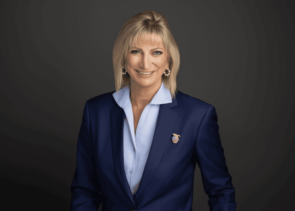 A person with short blonde hair wearing a blue blazer, light blue shirt, and a pin, posing and smiling in front of a dark background.