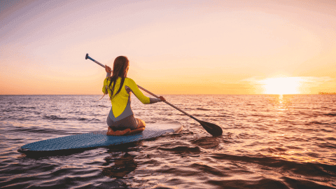 A person kneels on a Pro Balance 360 paddleboard, paddling on the ocean during sunset under a clear sky.