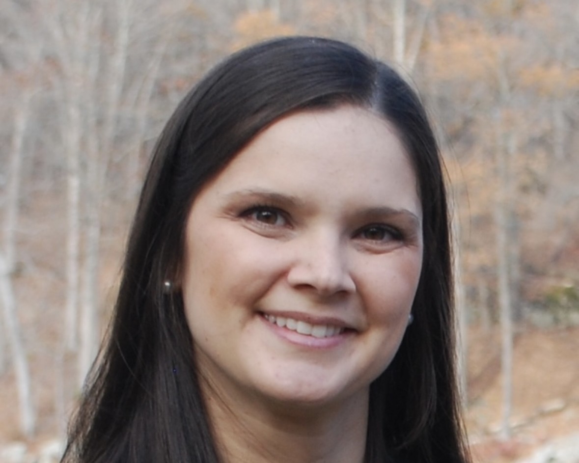 A woman with long dark brown hair smiles outdoors with autumn trees in the background.