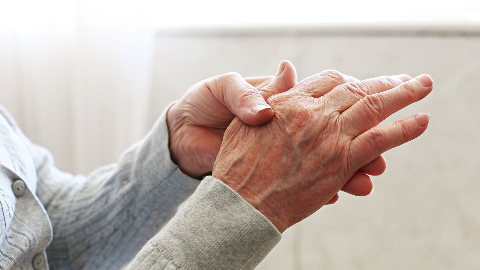 Close-up of an elderly person wearing a grey sweater, holding and rubbing their hand, possibly indicating joint pain or discomfort from arthritis.