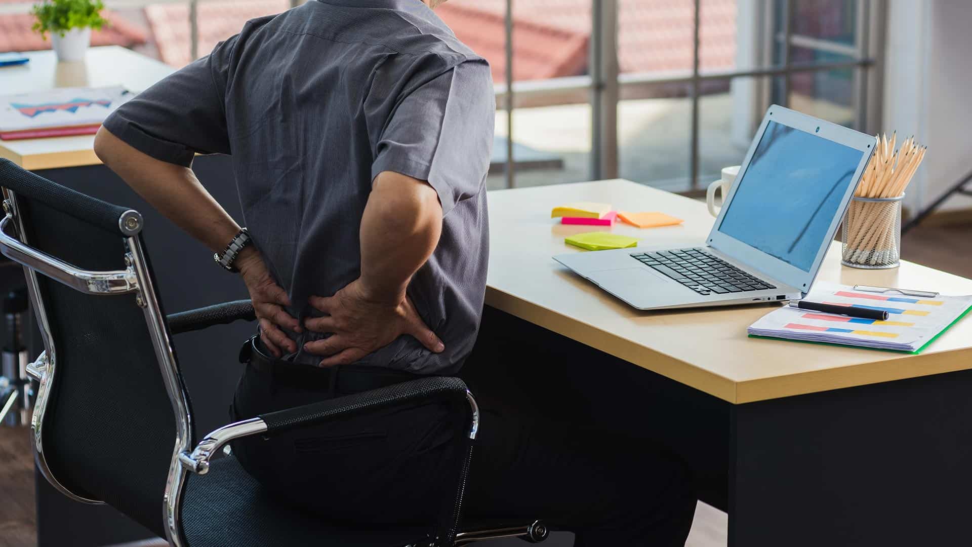 A person sitting at a desk in front of a laptop holds their lower back, appearing to experience back and neck pain in an office setting.