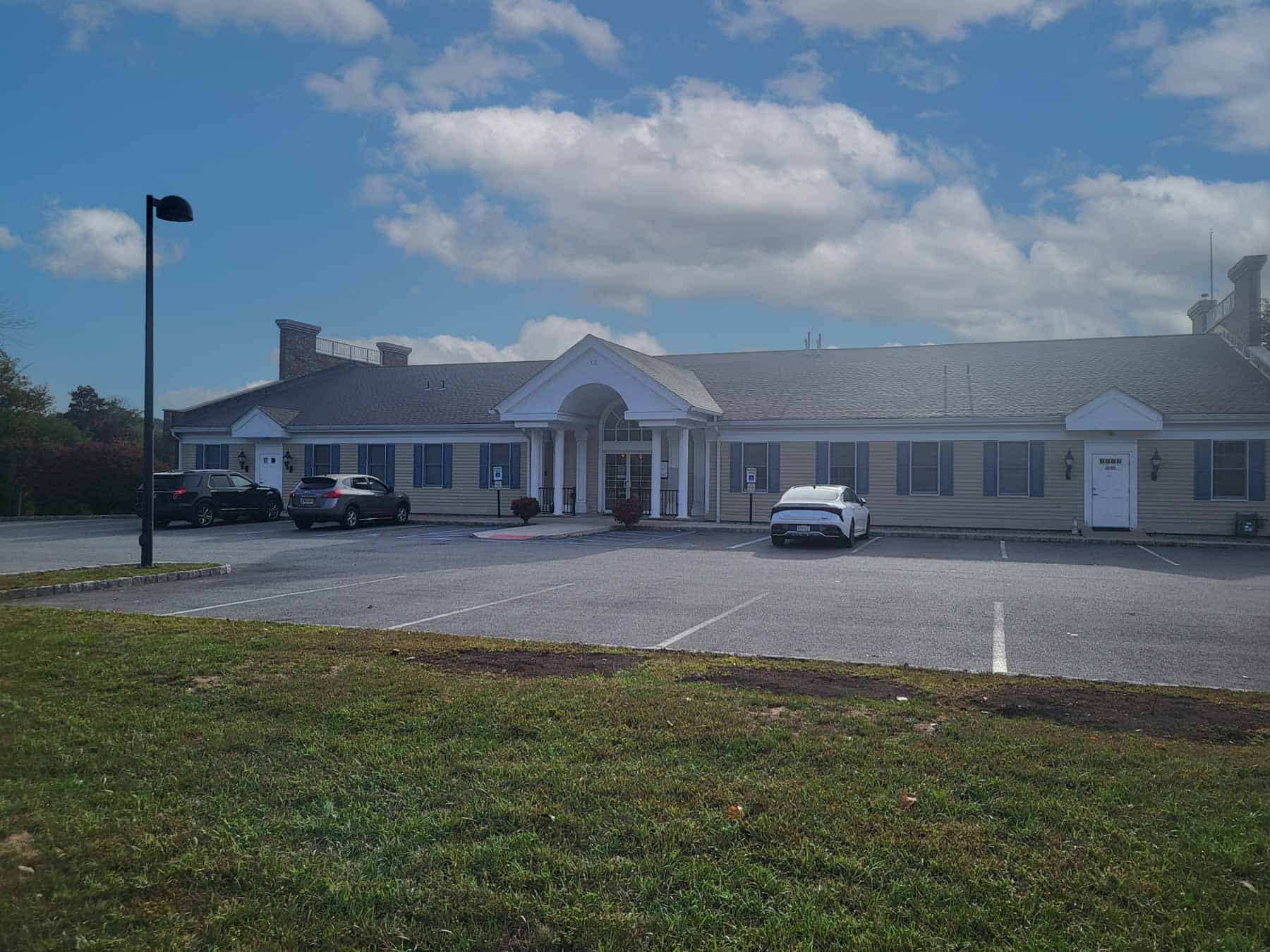 Single-story office building with white columns and a central entrance, three cars parked in front, and mostly empty parking spaces under a partly cloudy sky.