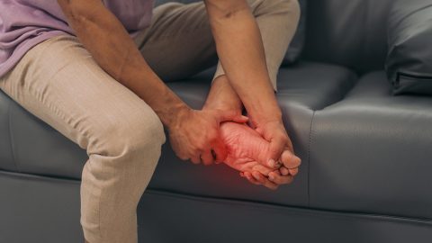 Person sitting on a couch holding their foot, with the sole highlighted in red to indicate pain or discomfort, a common sign of neuropathy.