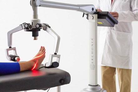 A medical professional operates a laser therapy device targeting a patient's foot, which is resting on an examination table.