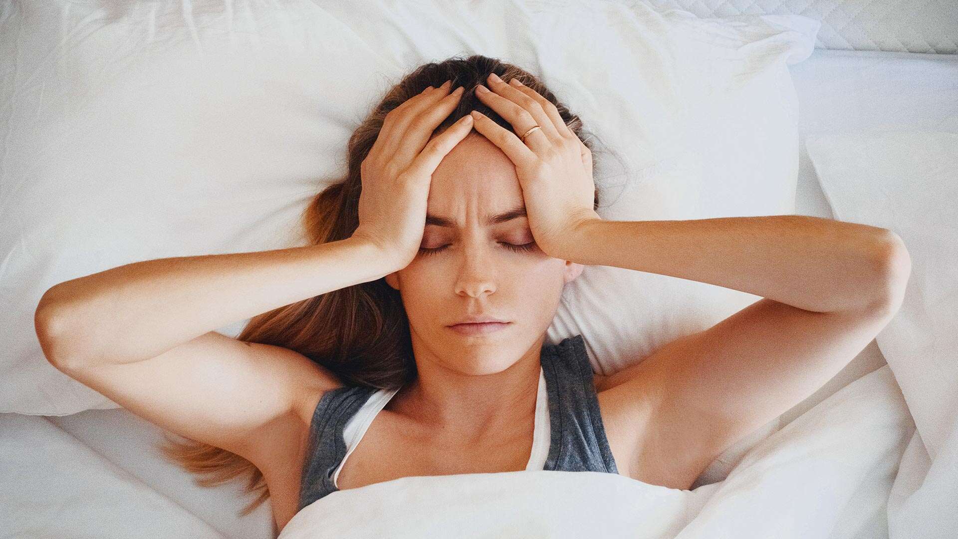 A woman lying in bed with her eyes closed, holding her head with both hands, appears to be experiencing discomfort or migraines.