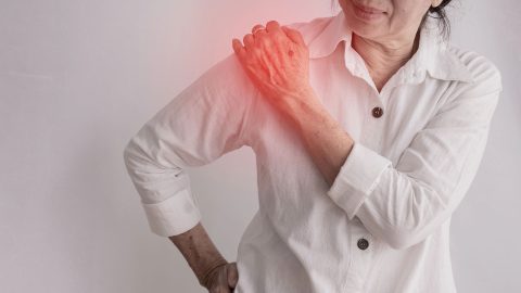 An older woman in a white shirt holds her left shoulder, which is highlighted in red, suggesting pain or discomfort and the possible need for pain management.