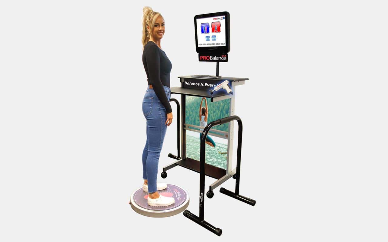 A woman stands on a circular balance platform connected to a machine with handrails and a screen displaying balance-related information.