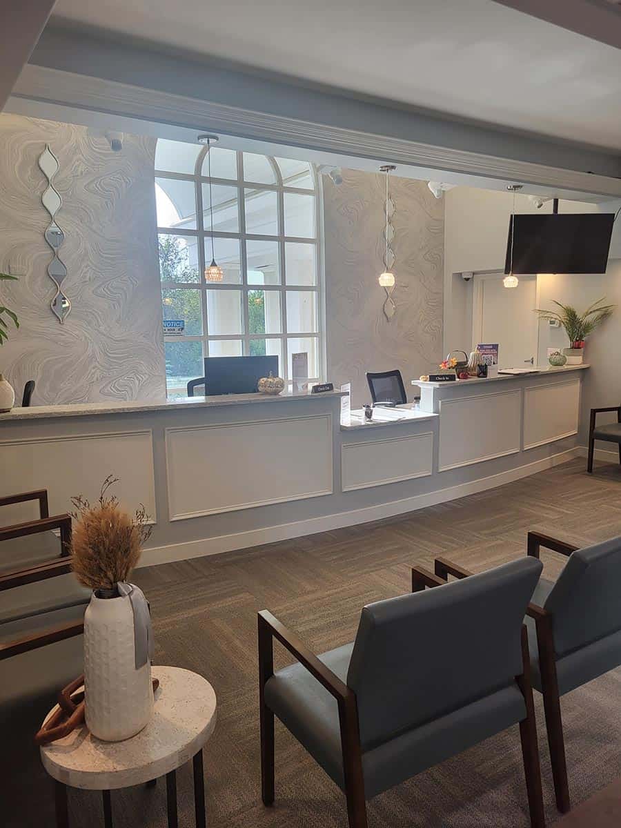 Modern waiting area with several chairs, a small table with a vase, and a reception desk featuring computers, decor, and a wall-mounted TV. Natural light enters through large windows.