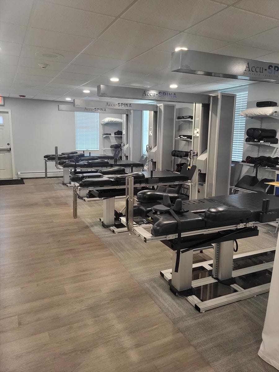 A row of four black medical treatment tables, labeled Accu-SPINA, are arranged in a clean, well-lit clinic room with wood flooring and white walls.