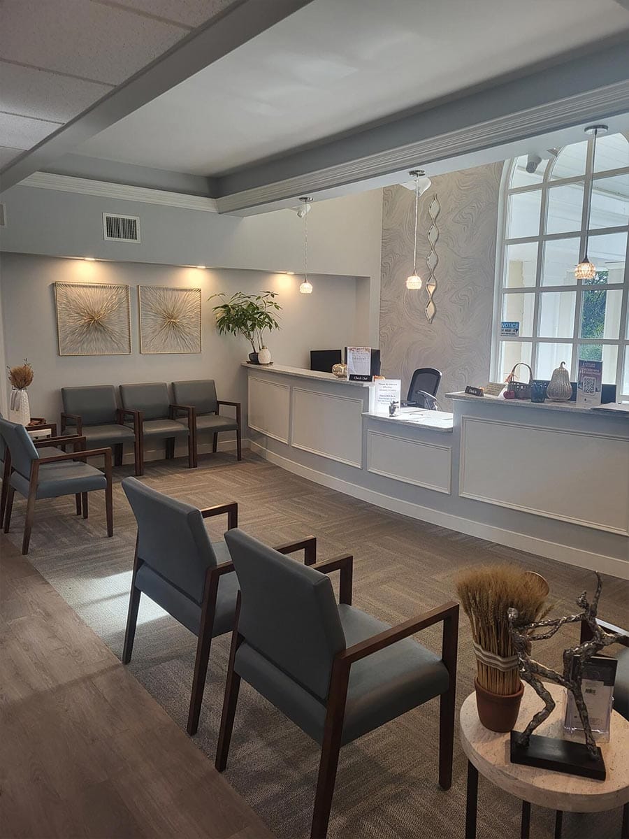 A modern waiting room with gray chairs, a reception desk, wall art, decorative plants, and large window letting in natural light.