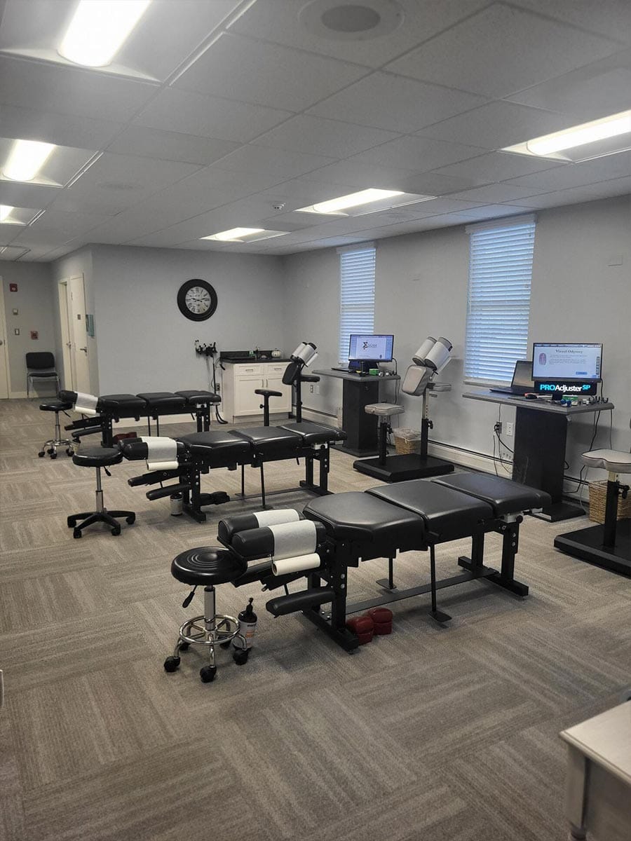 A clean, well-lit clinic room with three adjustable exam tables, stools, two computer stations, and medical equipment against white walls with windows and a wall clock.