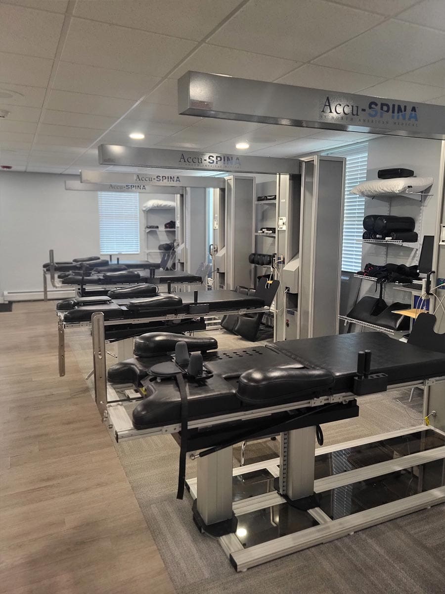 A row of Accu-SPINA spinal decompression machines in a well-lit, modern clinic room with windows and shelves holding equipment.