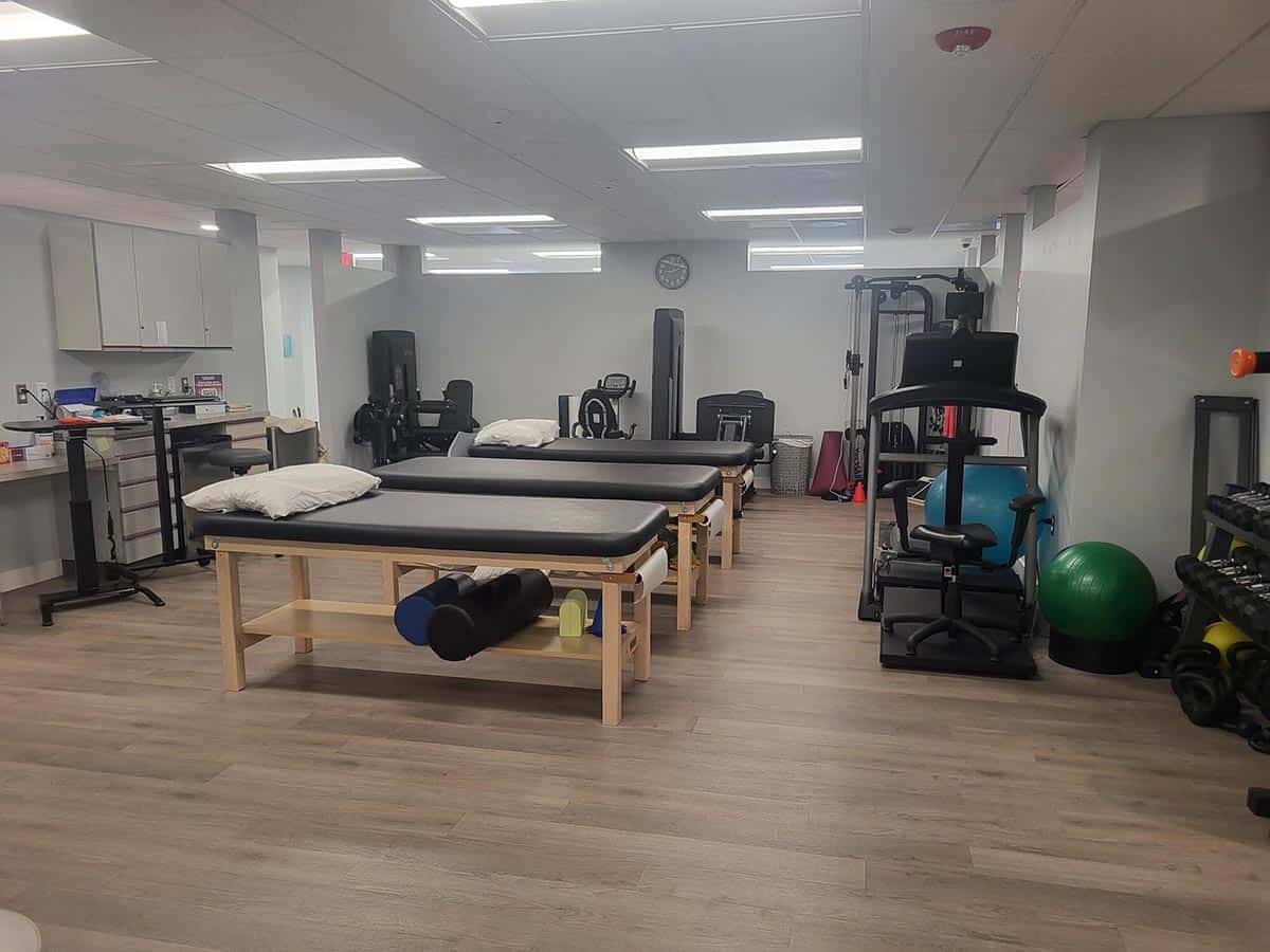 A physical therapy room with two treatment tables, exercise equipment, weights, and a therapy ball on a wooden floor.