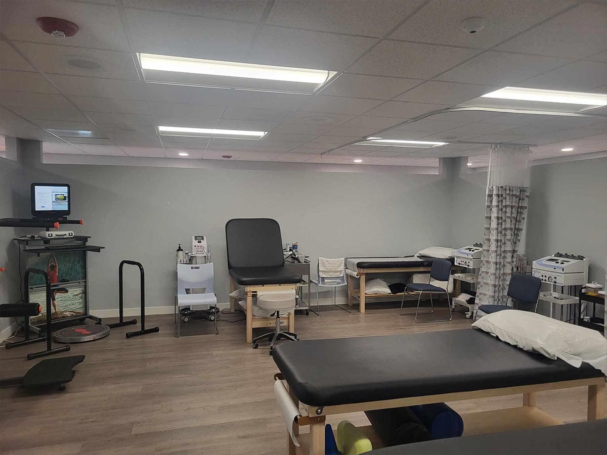 A physical therapy clinic room with treatment tables, exercise equipment, chairs, medical devices, and bright overhead lighting.