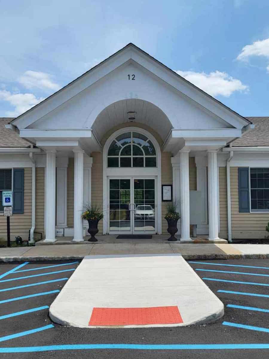 Single-story building entrance with white columns, arched window above glass doors, accessible ramp, blue sky, and marked parking spaces in front.