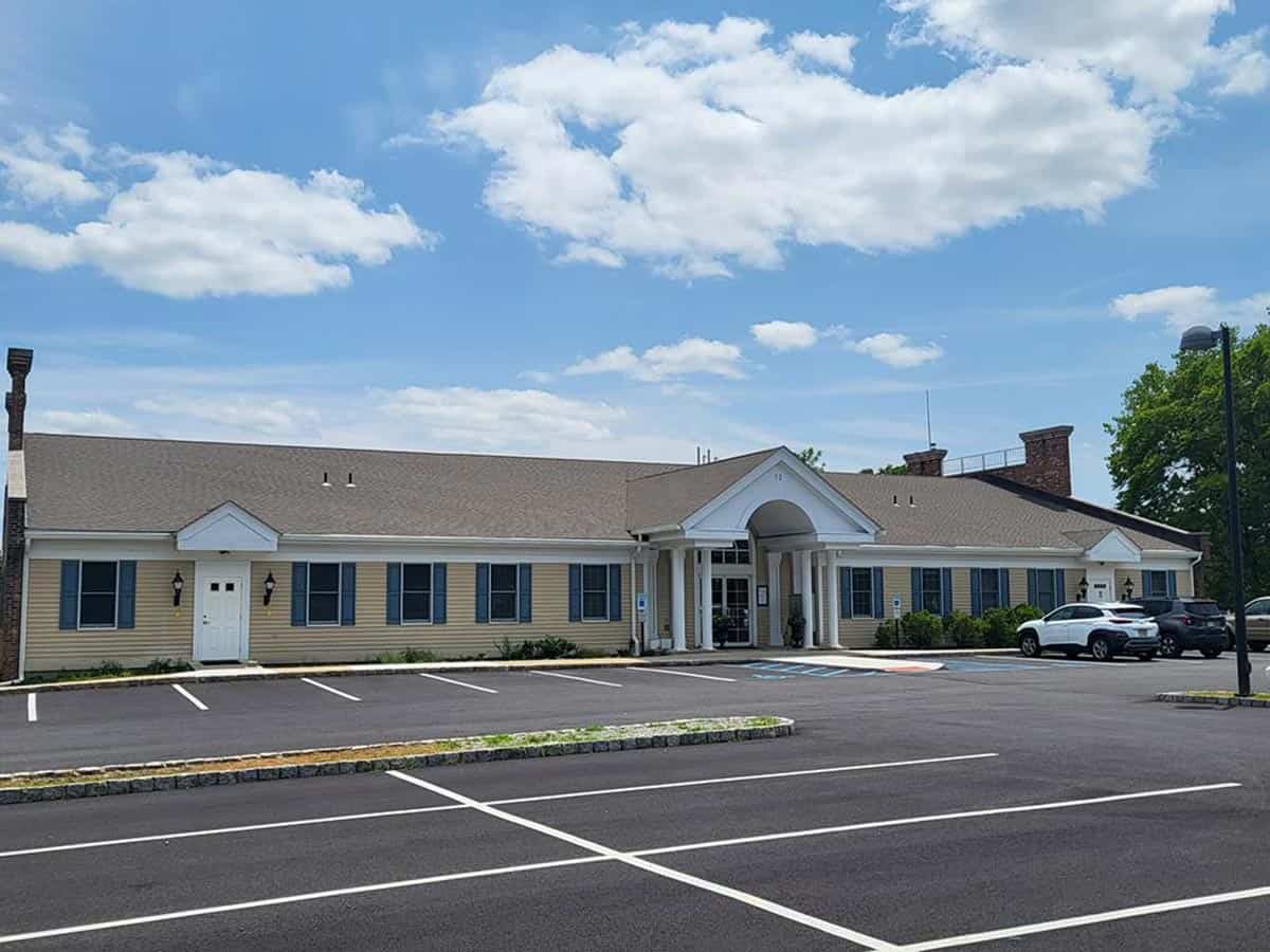 Single-story office building with beige siding, white trim, and a columned entrance; several cars parked, mostly empty parking lot, blue sky with scattered clouds.