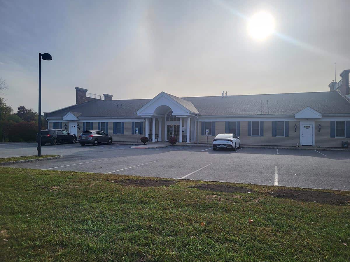 A single-story building with white columns and a parking lot in front, three cars parked, and the sun shining brightly in the sky.