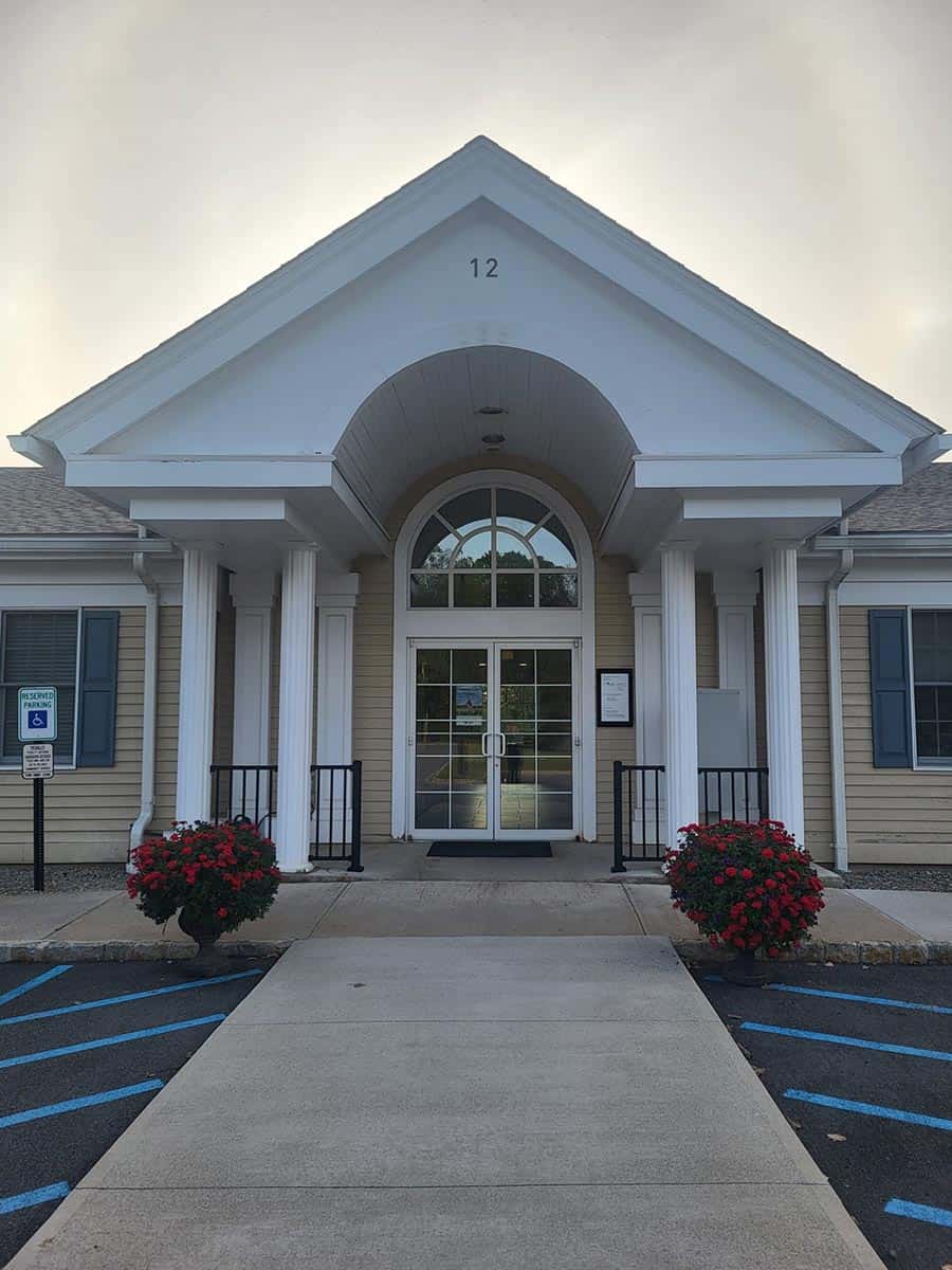 Single-story building with a white columned entrance, glass double doors, and two red flower bushes by the walkway. Handicap parking space and accessibility signs are visible.