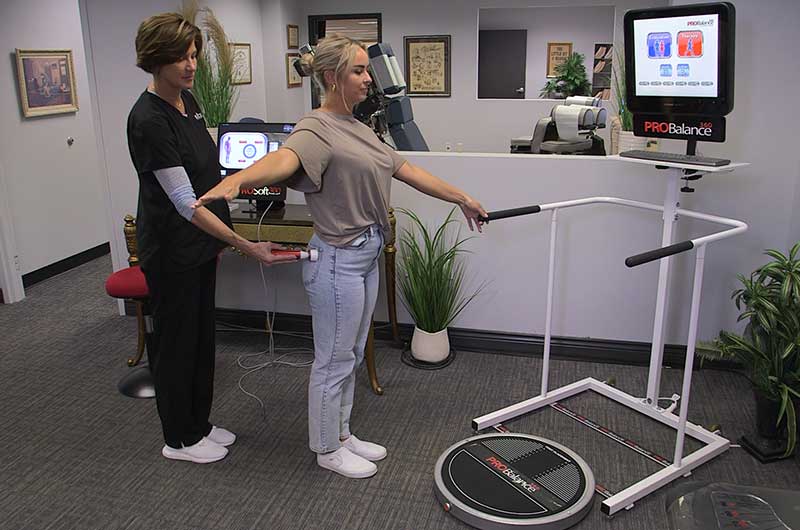 A woman stands with arms outstretched on a balance platform while a medical professional observes. Medical equipment and plants are visible in the room.