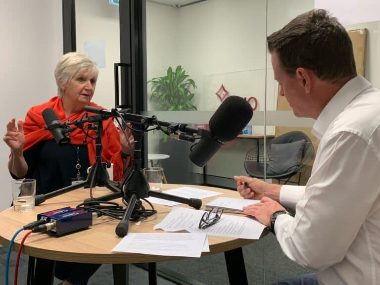 Recording session in an office meeting room, showing a woman in a red shawl speaking from across the desk, the host is wearing a white shirt and is facing her with his back to the camera.