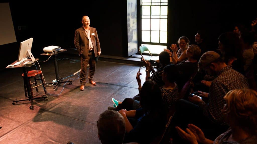 Martin Franklin standing in front of applauding crowd in small theatre. Light from a tall window on right shines across the floor
