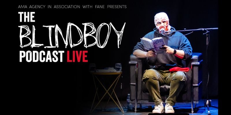 Blindboy Podcast Live. Image of podcaster, Blindboy sitting in an armchair speaking into a microphone holding a book. He has a plastic bag on his head with eye and mouth holes cut out.