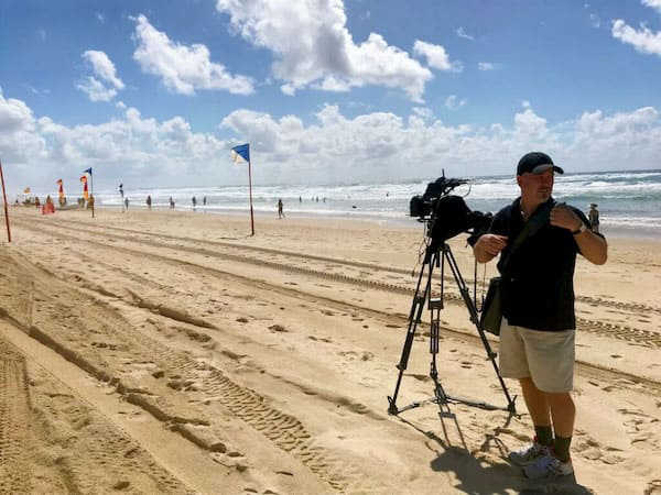 Filming on Broad Beach, Gold Coast, Commonwealth Games for the production of a documentation video for artist, Andrew Baines. Martin is wearing shorts and a baseball cap standing beside a large tripod and video camera.