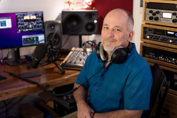 Podcast producer, Martin Franklin in front of a large desk, with audio speakers behind him and a rack of audio equipment to his left. He has headphones around his neck and is wearing a turquoise short sleeved shirt