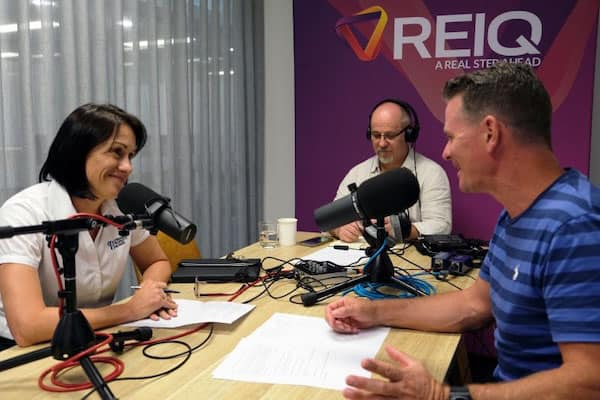 Recording for REIQ in office. Podcast Producer, Martin Franklin is wearing headphones looking down at a Sound Devices audio recorder. He is sitting under a purple banner with the REIQ logo, in front of him is podcast presenter, Rob Doorey interviewing a  guest with large microphones on stands