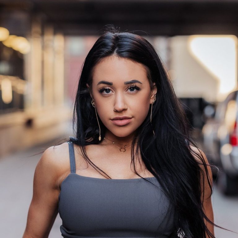 Portrait Of Paige Michael, Rn Standing In A City Alleyway, Wearing A Gray Tank Top And Gold Jewelry, Looking Confidently At The Camera With Long Dark Hair.