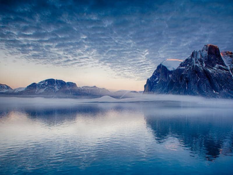 Scenic Greenlandic fjord with ice-capped mountains and unusual cloud formations reflecting in still blue waters