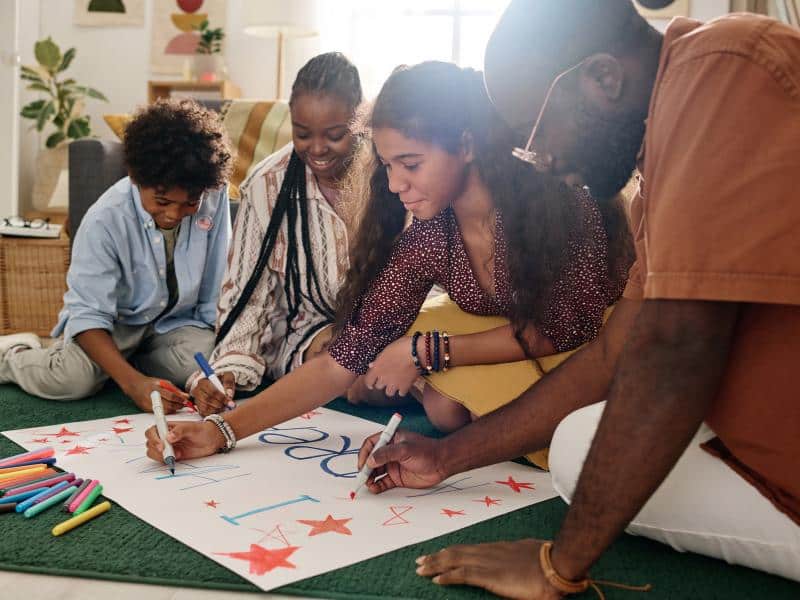 Group of people sitting on the floor collaborating on colorful poster-making project with markers and art supplies, focused on their creative work together.
