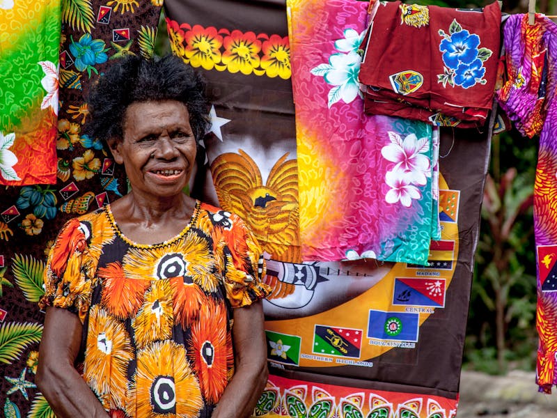 A senior woman stands smiling amidst vibrant traditional Papua New Guinean clothing.