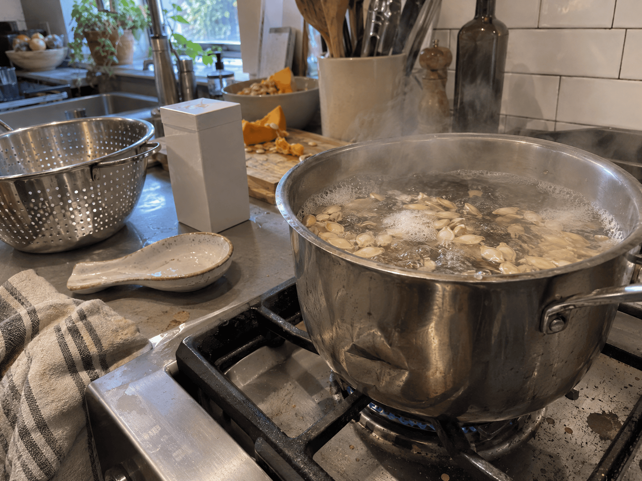Pumpkin seeds boiling in a pot of salted water on a stovetop with steam rising and a colander nearby