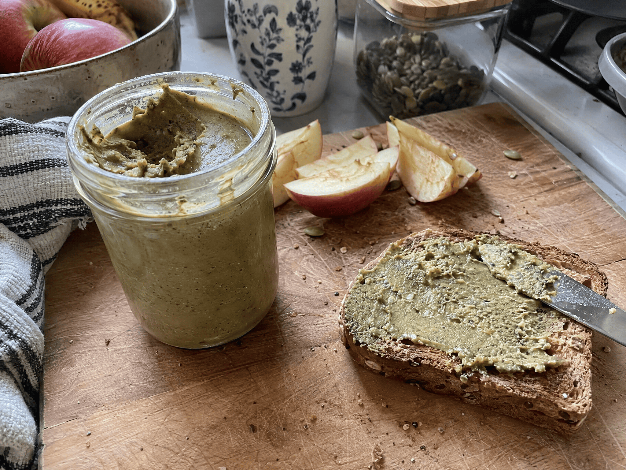 A jar of homemade pumpkin seed butter on a wooden cutting board with a knife on toast and apple slices nearby