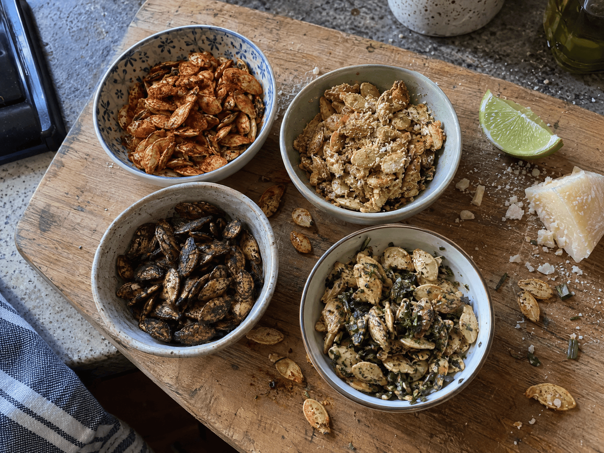 Four small bowls of different savory seasoned pumpkin seeds on a wooden board with a lime wedge and parmesan nearby
