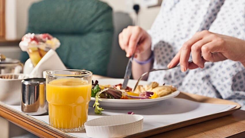 A table with orange liquid in a cup. A person uses a fork and knife on a plate on the table, The person wears a white robe with blue dots. There is a blue green chair in the background