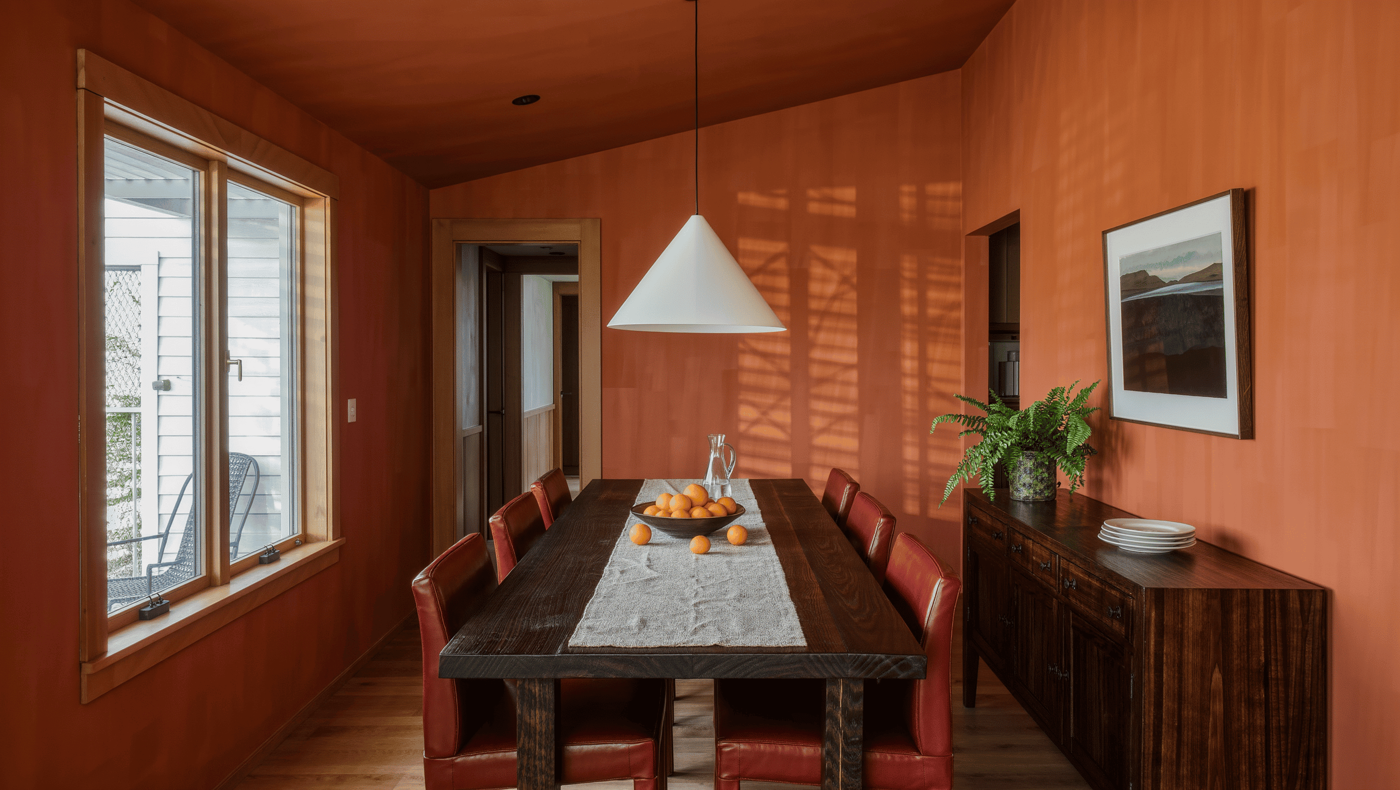A modest dining room dominated by a long dark oak table with oxblood leather chairs, leaving narrow passing space beside a slim sideboard.