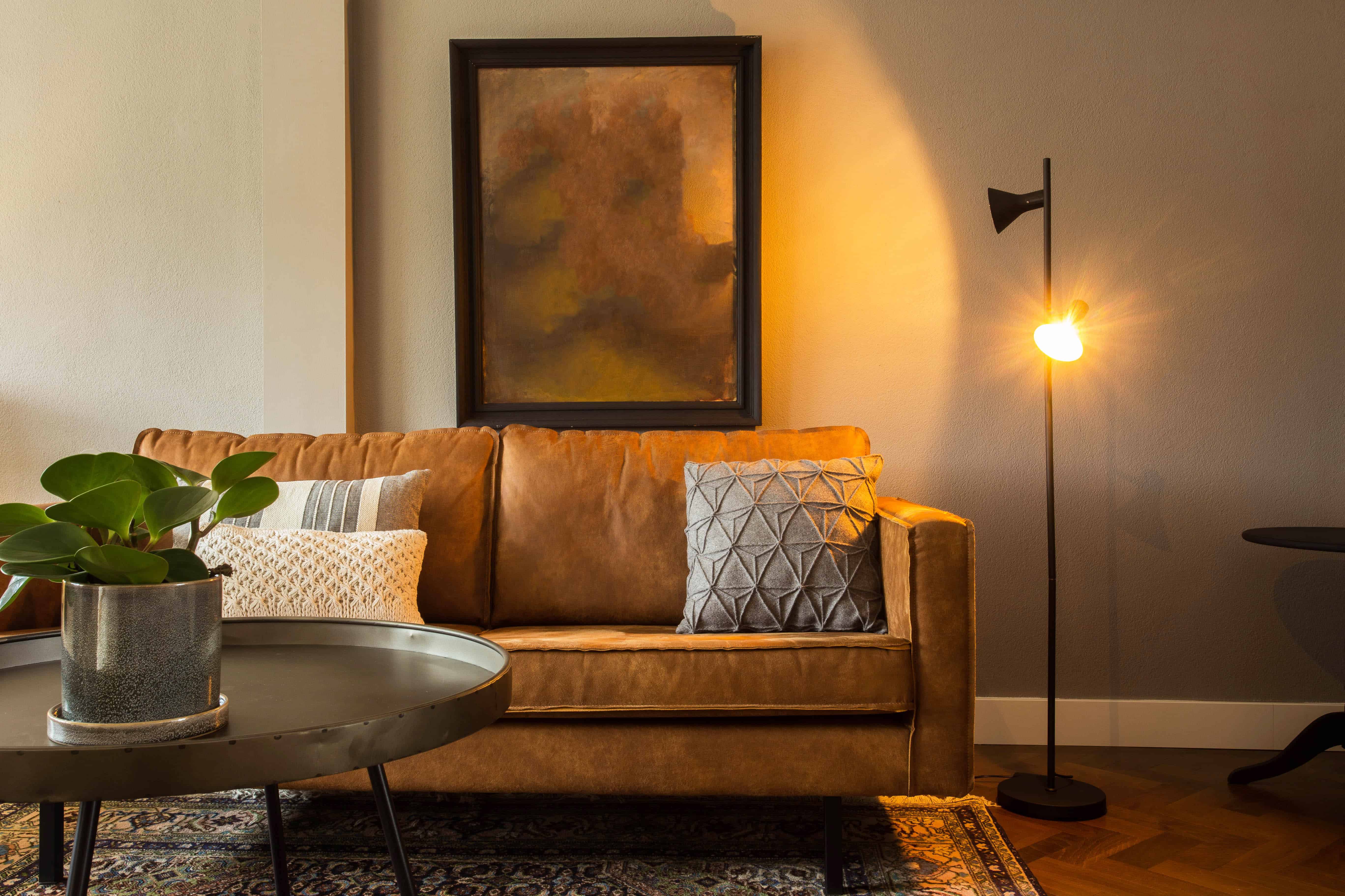A cognac leather sofa layered with mixed-pattern pillows sits beside a small round accent table, framed art, and a brass floor lamp on a patterned rug.